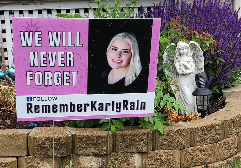 A We Will Never Forget yard sign with Karly's photo beside an angel statue in a garden.