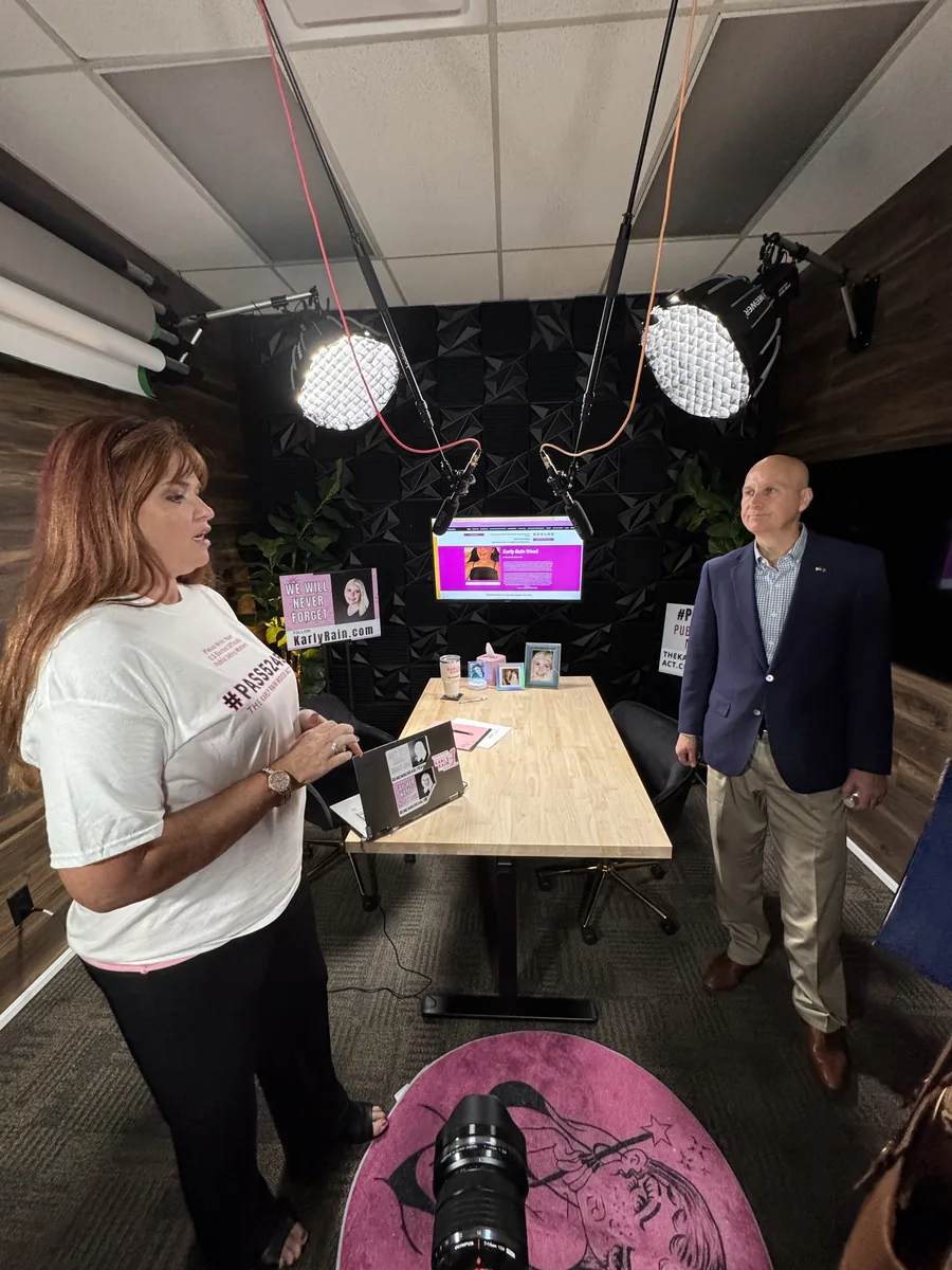 Amber Wood talking with Senator Pete Ricketts in a podcast studio, with Karly's page on the screen.